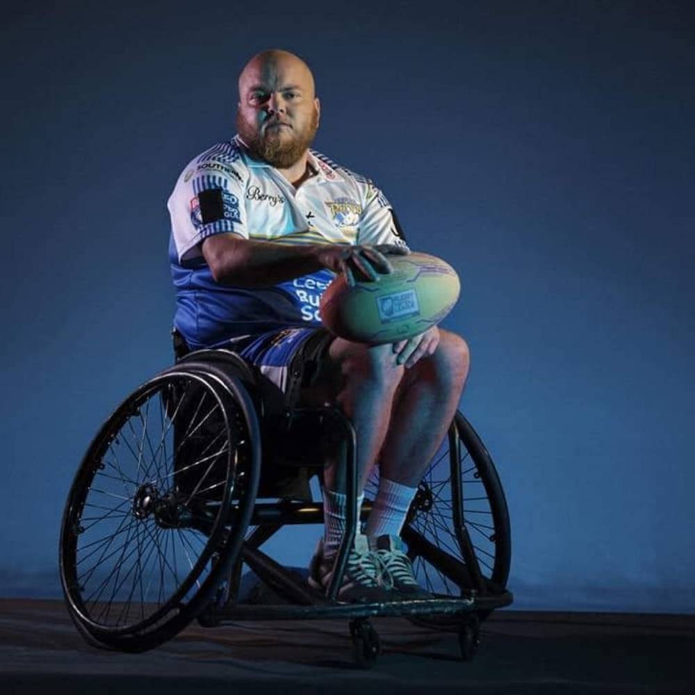 Verity Smith, a rugby player, wears a white uniform as he sits in a wheelchair while holding a rugby ball on his lap
